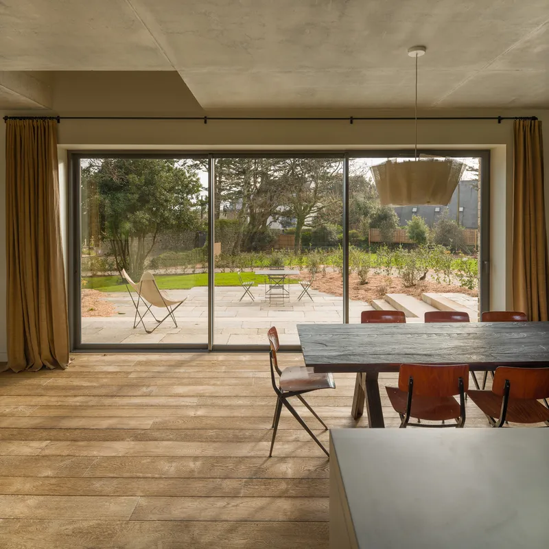 Dining room with full-height glazing overlooking garden terrace with outdoor chairs and lawn beyond
