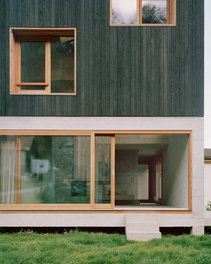 Facade detail showing timber window frames set in dark vertical cladding and white concrete base