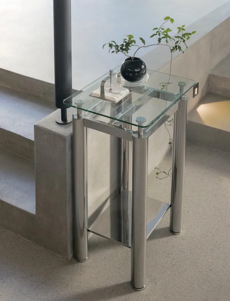 Glass and steel side table holding a black vase with green foliage beside concrete steps