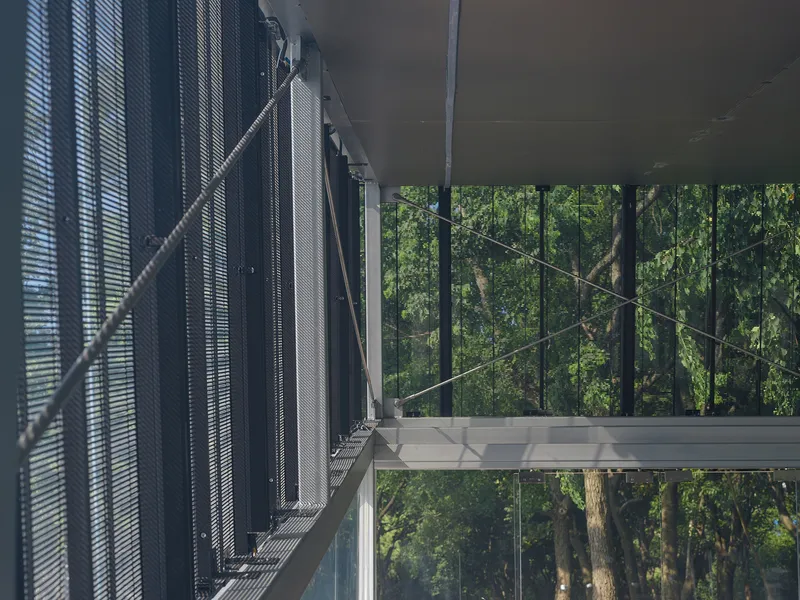Interior view through perforated metal screens toward floor-to-ceiling glass overlooking dense forest