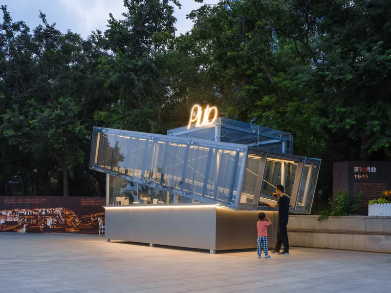 Illuminated glass kiosk with angled canopy and neon signage at dusk with two visitors at the counter
