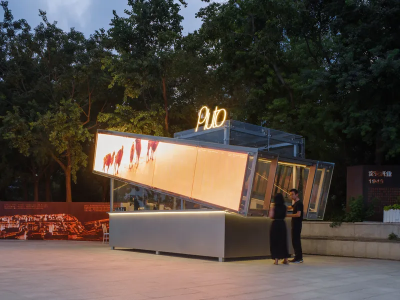 Glass pavilion with backlit panels and neon sign at twilight with a visitor leaning against the counter