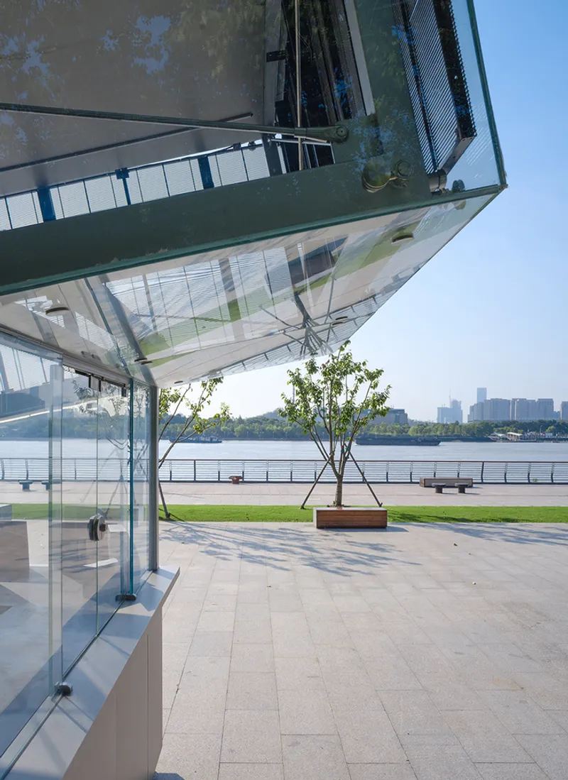 Cantilevered glass volume above a waterfront plaza with a single planted tree in a timber planter