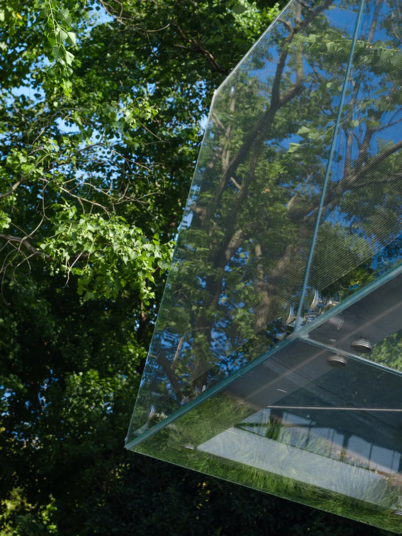 Angled glass facade reflecting surrounding tree canopy against a summer sky
