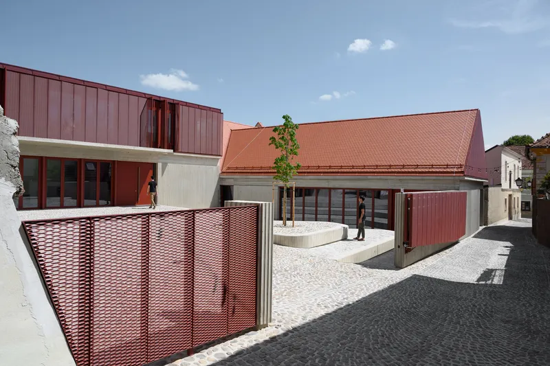 Cobblestone courtyard showing perforated red metal screen fence and sloped tile roof under blue sky