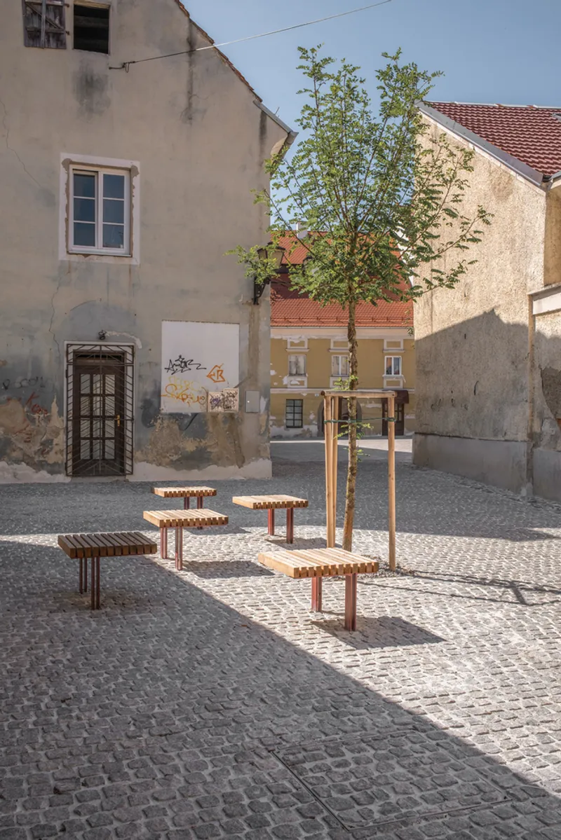 Cobblestone plaza with timber and metal seating elements around a young tree and weathered stucco facades
