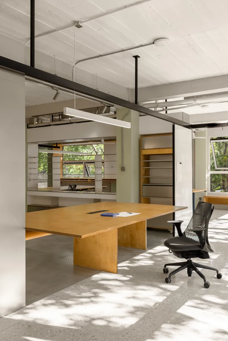 Double-height workspace interior with exposed ceiling joists and plywood desk lit by dappled sunlight