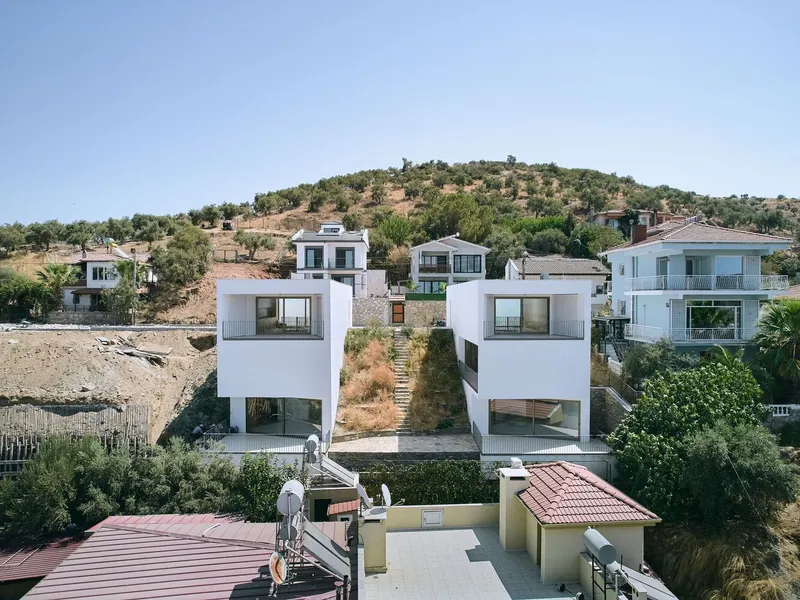 Aerial view of two white cubic volumes on a hillside lot among neighboring houses under clear sky