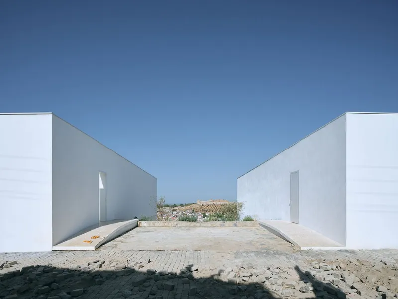 Two white volumes frame a cobblestone courtyard with distant farmhouses visible under a clear blue sky