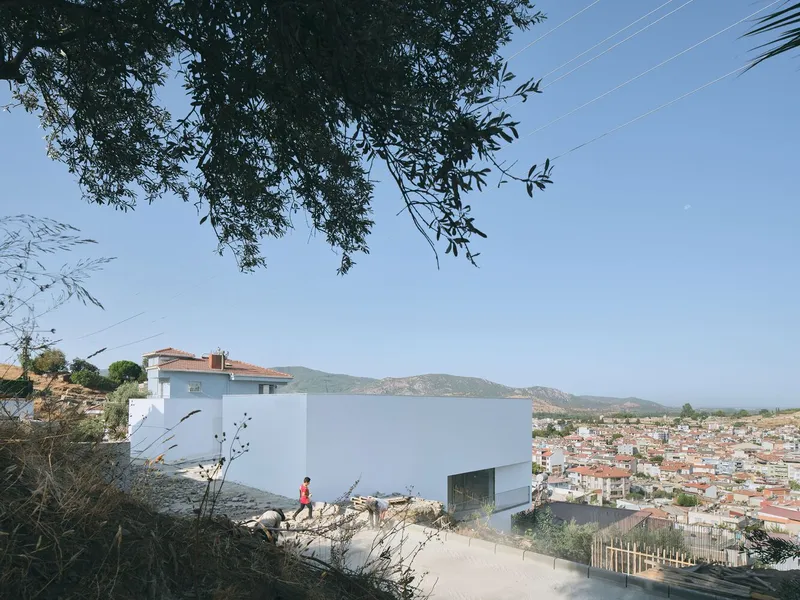 White plastered volume set into sloped terrain with person in red walking beneath olive tree canopy