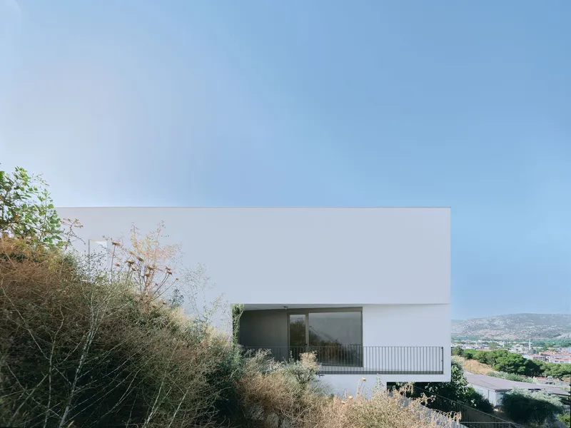 White rendered facade with cantilevered volume and recessed glazing surrounded by dry summer vegetation