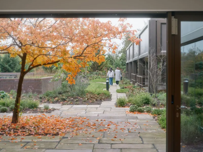 View through glass doors to stone-paved garden path with two figures walking beneath autumn foliage