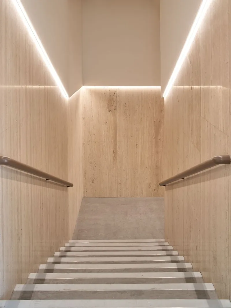 Travertine staircase descending between cove-lit walls with bronze handrails on either side