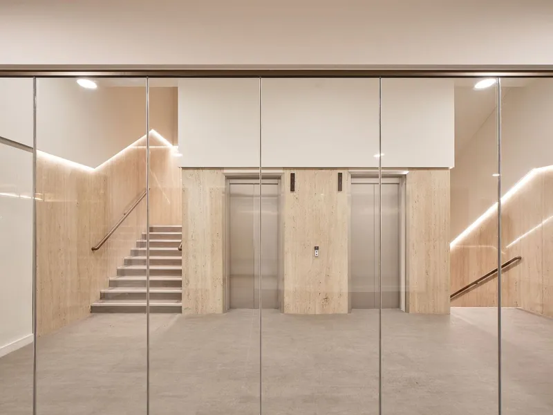 Glass-walled lobby with travertine elevator surrounds and cantilevered stairs under recessed lighting