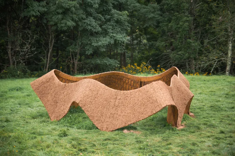 Undulating cork pavilion with ribbed interior ceiling sitting in a green meadow edged by forest