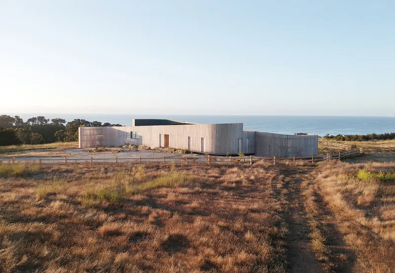 Weathered timber-clad residence set in dry coastal grassland with the ocean visible beyond