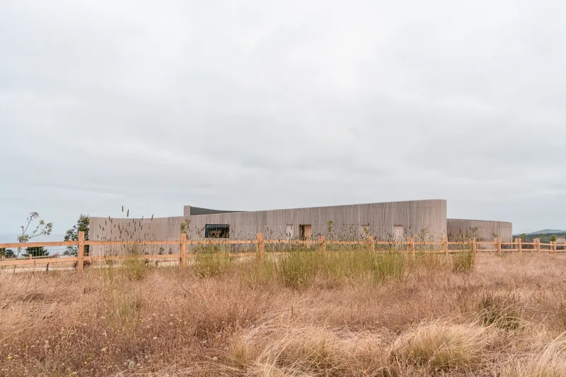 Horizontal timber-clad facade viewed across a field of dry grasses under overcast skies
