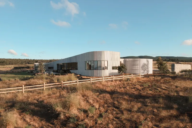 Curved timber-clad volumes connected by glazed sections behind a timber post fence