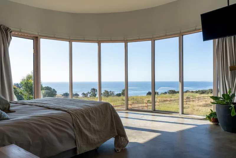Curved glazed wall in bedroom framing ocean and coastal grassland views under clear sky