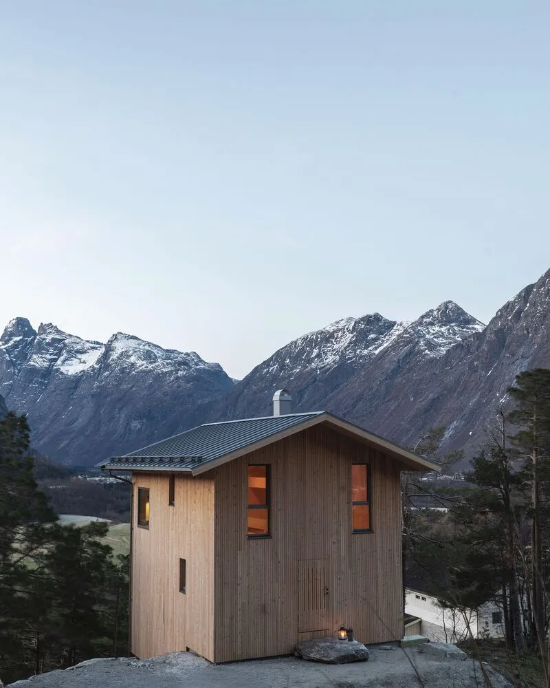 Vertical timber facade with illuminated windows at twilight framed by evergreen trees and distant peaks