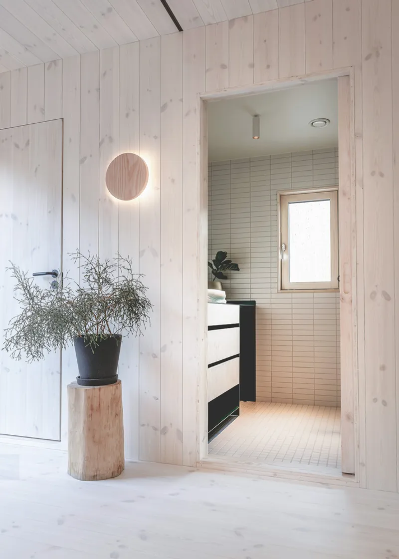 Interior hallway with circular wall light and potted plant leading to bathroom with horizontal tile