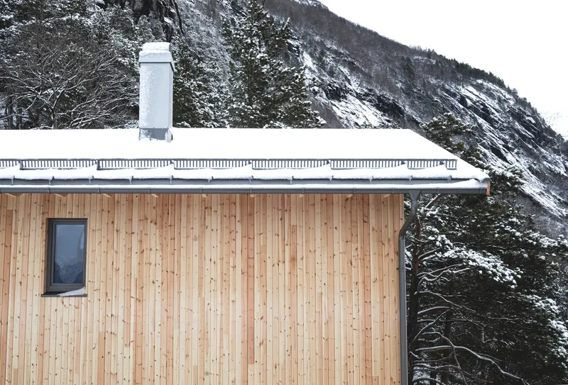 Snow covered gable roof with chimney against steep forested mountain slope in winter
