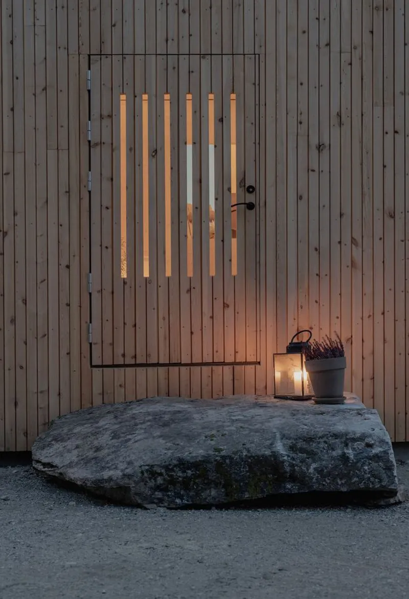 Timber clad entrance door with vertical slot windows and rock slab threshold at dusk