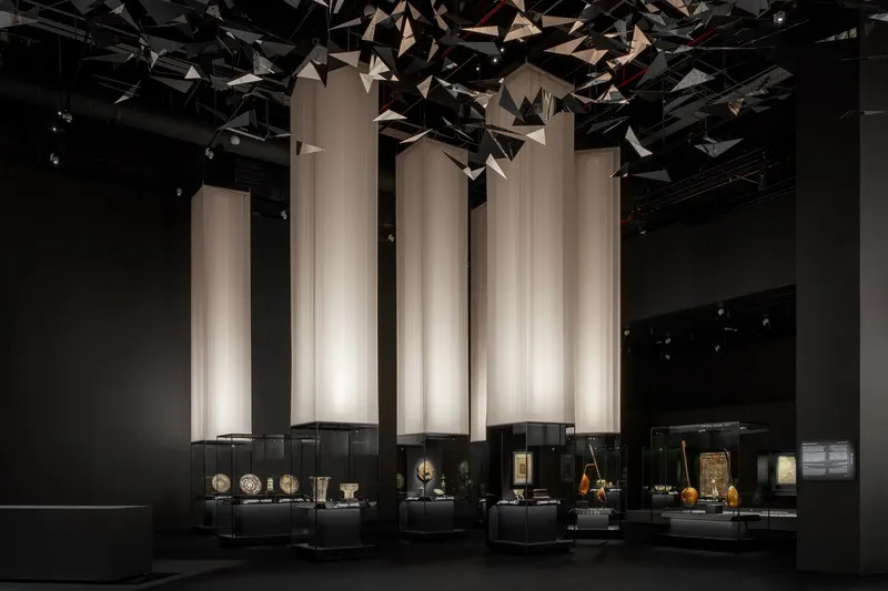 Gallery hall with illuminated vertical fabric screens above glass display cases beneath a faceted ceiling installation