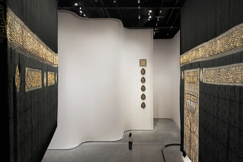 Gallery corridor between dark walls with inset gilded panels leading to a white curved partition wall