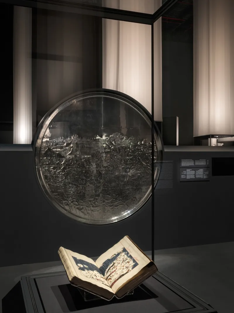 Close-up of circular engraved metal plate and open manuscript displayed within a glass vitrine