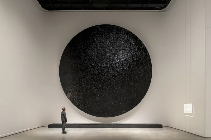 Large circular black textured wall installation with a lone visitor standing before a low horizontal platform