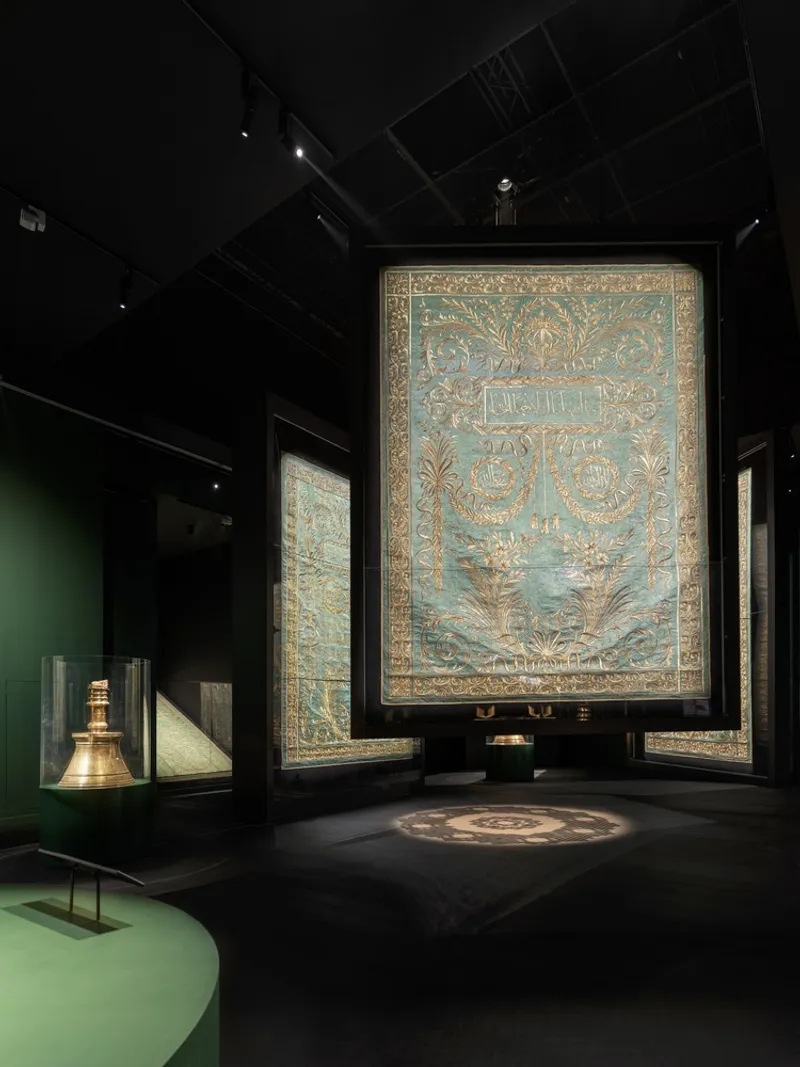 Blue and gold textile hanging in a black gallery with brass lantern in vitrine and projected floor pattern