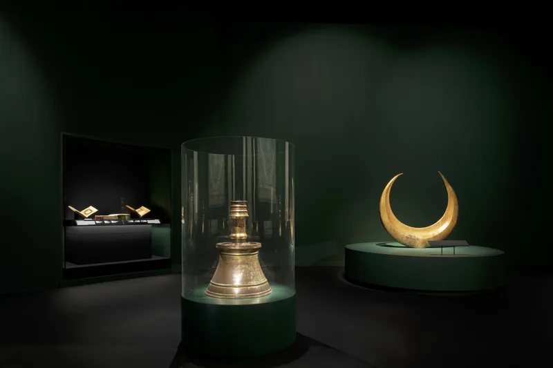 Gallery display with cylindrical glass cases on green plinths containing artifacts under focused spotlighting