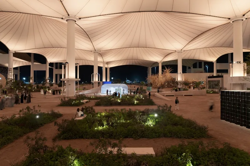 Plaza with landscaped beds and red clay paving beneath tensile canopies at night with illuminated plantings