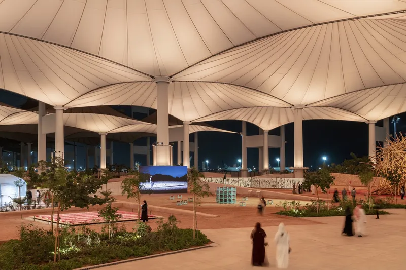 Tensile fabric canopy with radiating pleats supported by white columns at dusk with visitors below