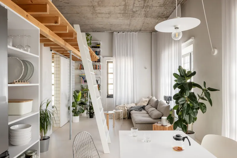 Open-plan interior with white ladder staircase ascending to timber loft under raw concrete ceiling