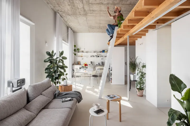 Double-height living space with exposed concrete ceiling and timber loft staircase under bright daylight
