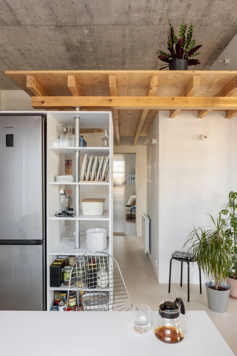 Kitchen view through open shelving unit showing timber-framed loft structure and concrete ceiling above