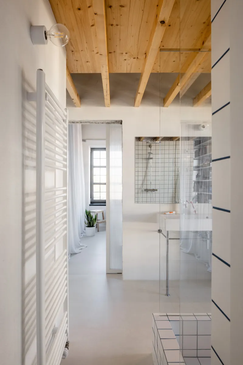 View through bathroom corridor showing white grid tile shower platform and exposed timber joist ceiling in natural light