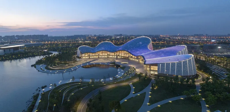 Three-lobed structure with undulating blue-lit roof rising above waterfront landscape at twilight