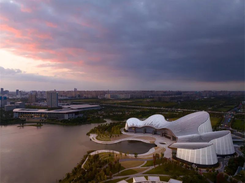 Aerial view of the curved ribbed roofline beside a lake and landscaped paths at sunset