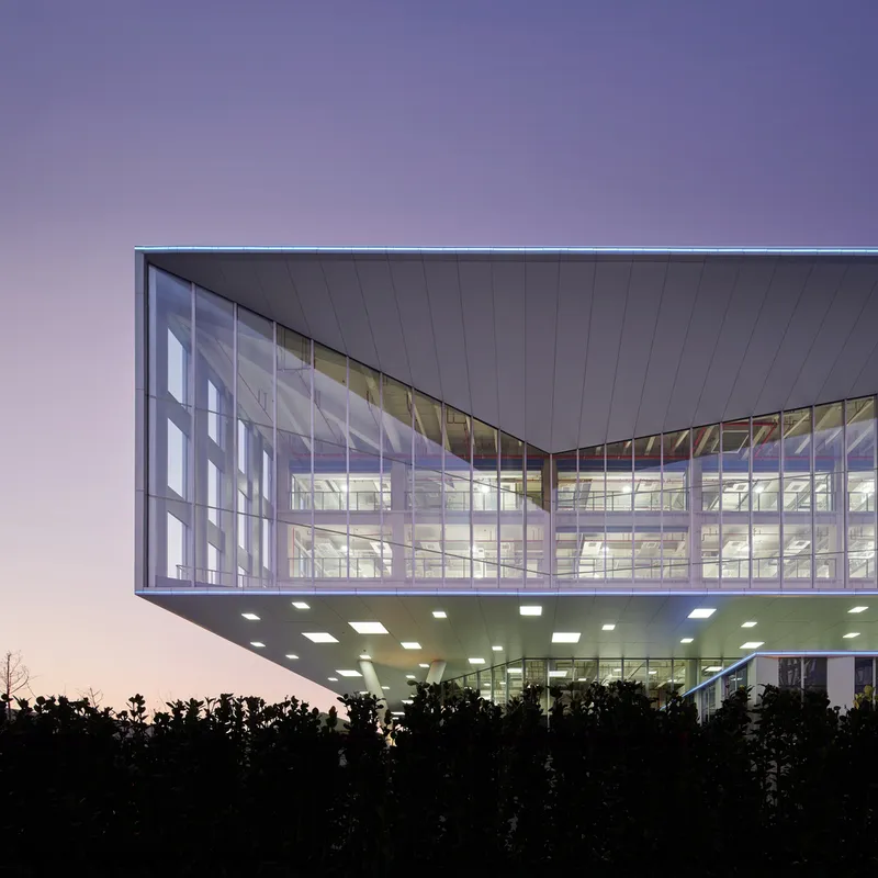 Cantilevered glass facade with illuminated interior visible above silhouetted trees at dusk