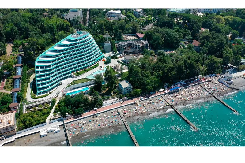 Aerial view of a curved glass facade hotel rising above a waterfront beach crowded with umbrellas