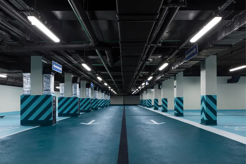 Long parking level with repeating columns and diagonal striped barriers beneath a black exposed structure ceiling