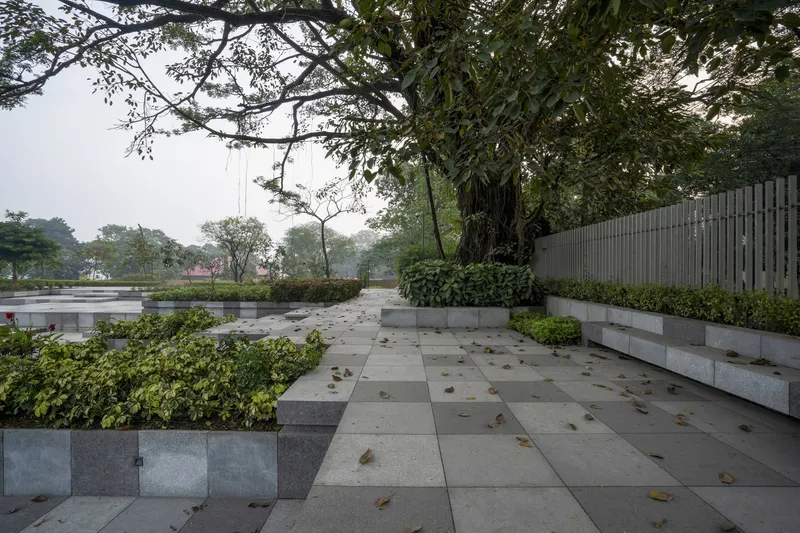 Paved courtyard with planted beds and fallen leaves beside a vertical slatted screen wall in hazy light