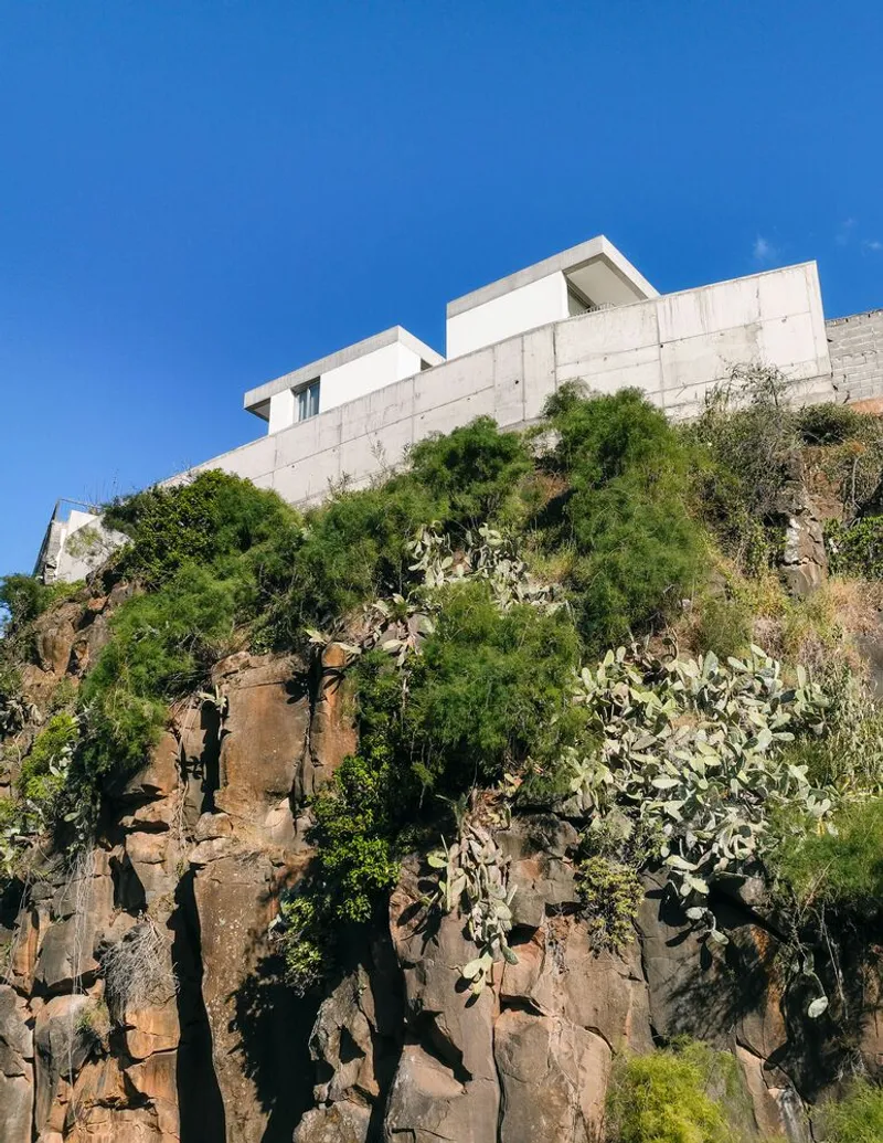 White concrete volumes perched atop a volcanic rock cliff with cacti and wild vegetation