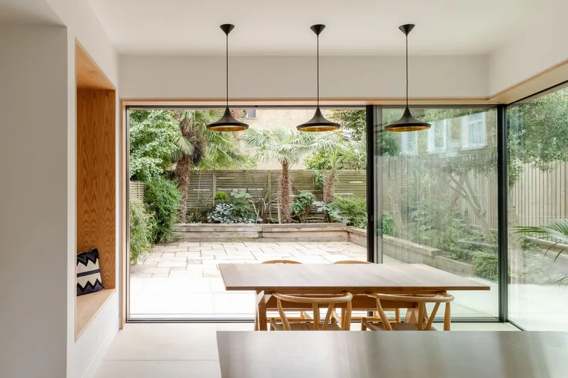 Dining area with pendant lights overlooking paved terrace planted with palms and ferns through glazed wall