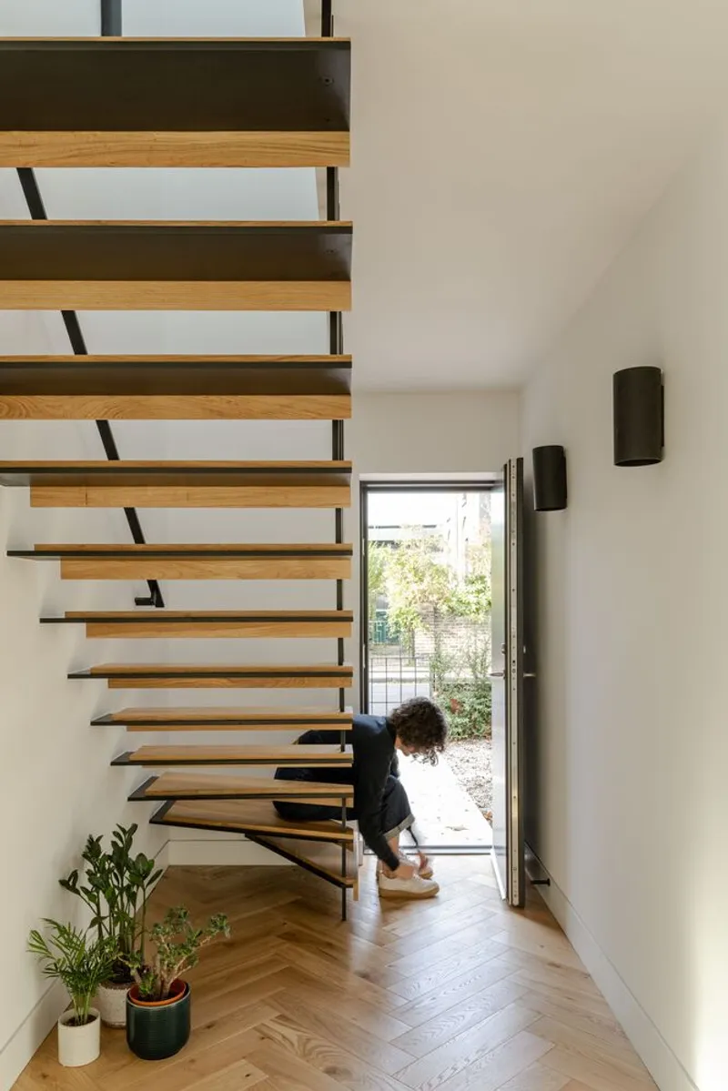 Cantilevered timber and steel staircase beside entrance door where a person tends to potted plants