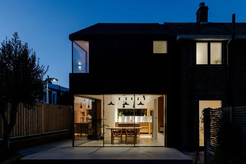 Night view of two-storey extension contrasting dark upper volume with lit ground-floor interior