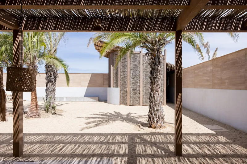 View through open timber pergola to woven screen pavilion framing palms and sand courtyard